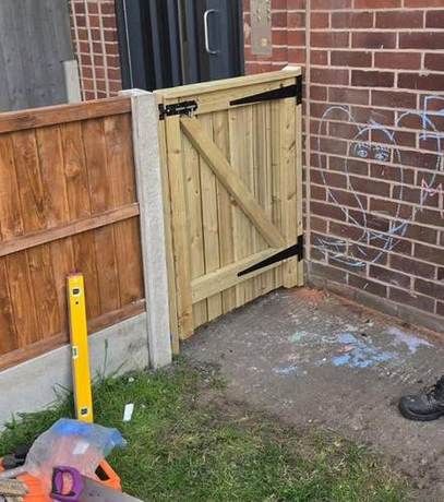 New timber gate hung on black strap hinges between a fence and a brick wall, with a spirit level alongside